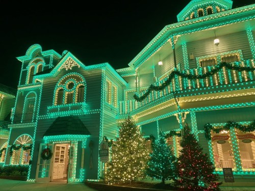 A beautifully lit house adorned with turquoise and white lights, surrounded by festive trees during the night.
