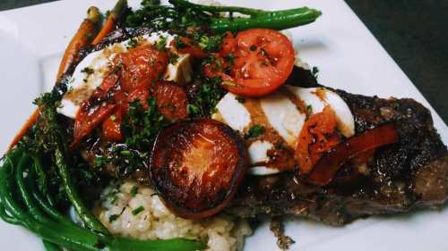 A plated dish featuring steak topped with tomatoes, peppers, and herbs, served with green beans and risotto.