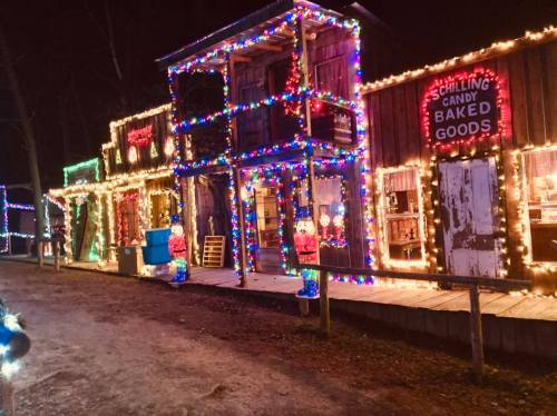 Colorful holiday lights adorn rustic storefronts in a festive nighttime scene.
