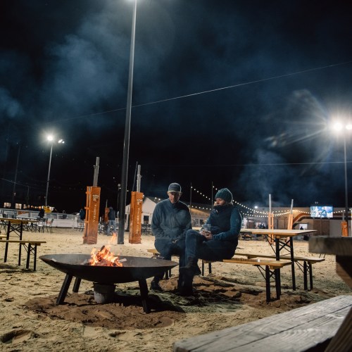 Two people sit by a campfire at night, surrounded by benches and string lights, with smoke rising in the cool air.