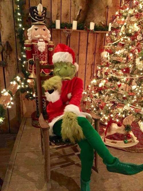 The Grinch in a Santa hat sits in a chair beside a nutcracker and a decorated Christmas tree.