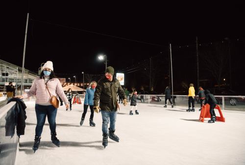 People ice skating at night, wearing masks, with festive lights in the background.