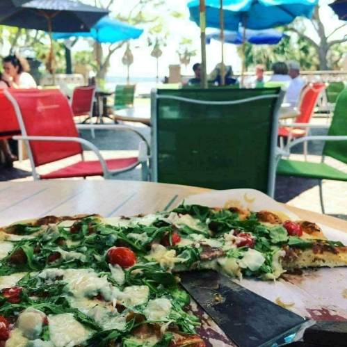 A close-up of a pizza slice with arugula and tomatoes, with colorful outdoor seating and a beach view in the background.