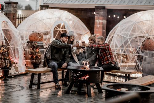 A group of four friends toasting drinks around a fire pit in a cozy outdoor setting with transparent igloos.
