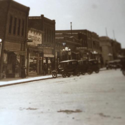 Historic street scene featuring vintage cars and early 20th-century buildings, with shops and a lunch counter visible.