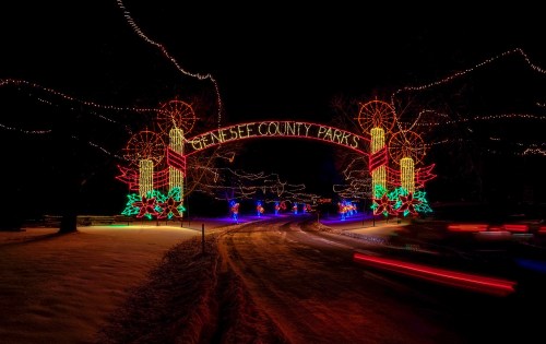 Colorful holiday lights illuminate the entrance to Genesee County Parks at night, with a snowy road and festive decorations.