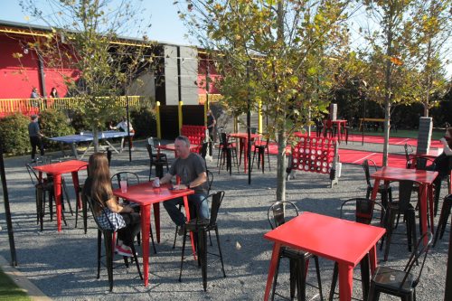 Outdoor seating area with red tables and chairs, surrounded by trees and a play area in the background.