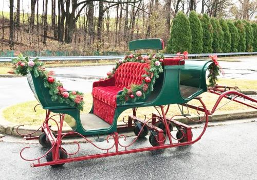 A festive green and red sleigh decorated with garlands and ornaments, parked on a road with trees in the background.