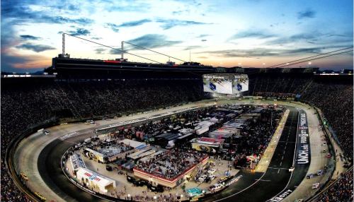 Aerial view of a bustling NASCAR race at a large stadium, with a vibrant sunset in the background.