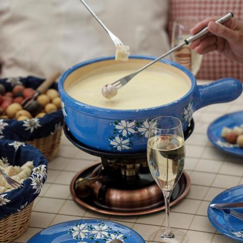 A blue fondue pot filled with melted cheese, surrounded by bread, fruit, and sparkling wine glasses on a table.