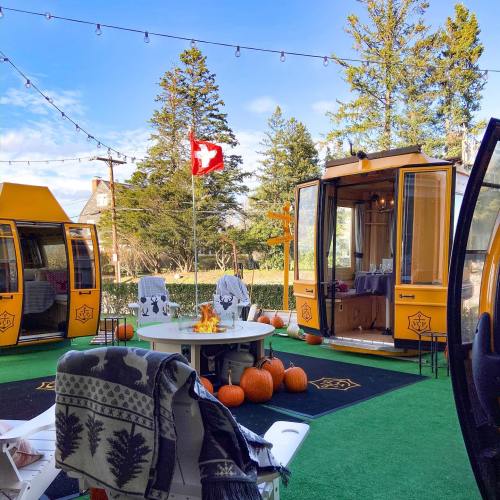 Cozy outdoor space with yellow cabins, a fire pit, pumpkins, and string lights, surrounded by trees and blue sky.