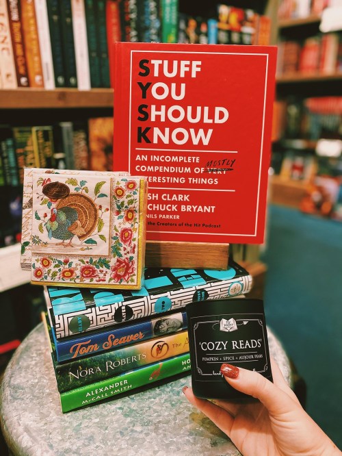 A hand holds a candle labeled "Cozy Reads" next to books and a decorative box on a table in a bookstore.