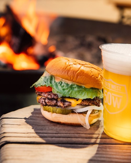 A juicy burger with lettuce, tomato, pickles, and cheese next to a frosty beer, with a fire in the background.