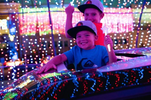 Two smiling boys in hats hold light wands, enjoying a colorful, illuminated display from a car window.