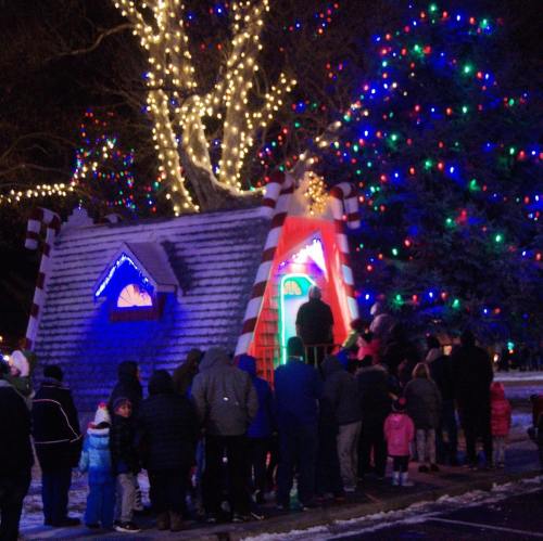 A crowd gathers in front of a brightly lit holiday display with a decorated tree and a whimsical candy-themed structure.