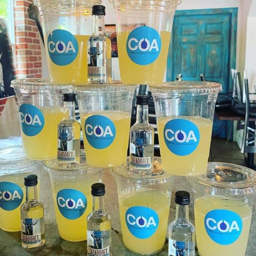 A stack of clear cups filled with yellow drinks, labeled with "COA," alongside small bottles of tequila.