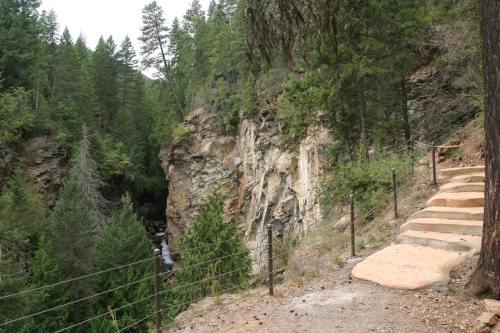 A winding path leads through a forested area with rocky cliffs and a small waterfall in the background.