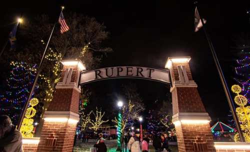 A festive entrance archway reading "Rupert," surrounded by colorful holiday lights and people enjoying the scene at night.