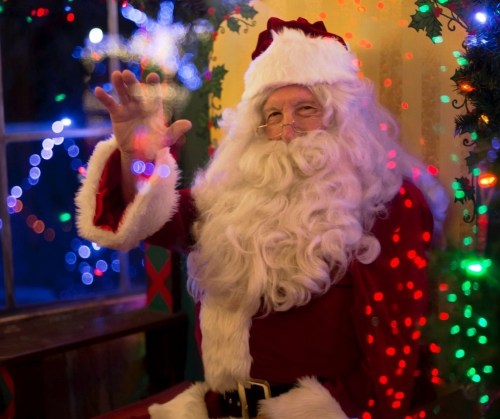 Santa Claus waves cheerfully, surrounded by colorful holiday lights and decorations.