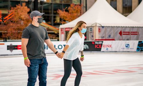 A couple ice skating together while holding hands, both wearing masks in an outdoor rink.