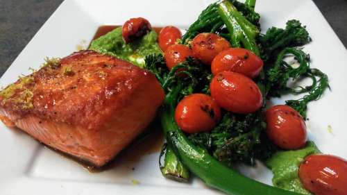 A plate featuring grilled salmon, cherry tomatoes, broccolini, and a green puree, garnished with lime zest.
