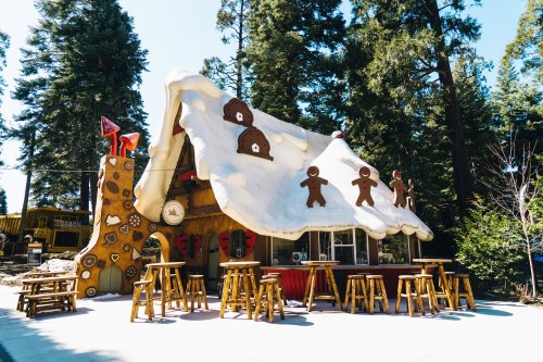 A whimsical gingerbread house with a snowy roof, surrounded by tall trees and wooden stools outside.