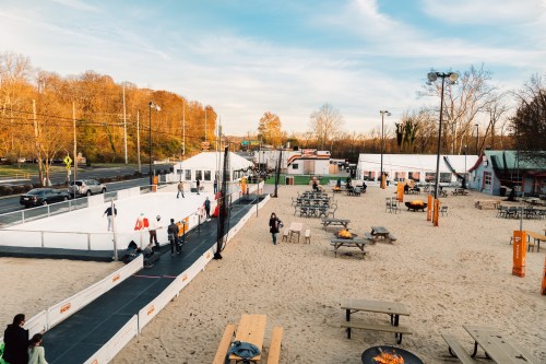 A sandy outdoor area with an ice skating rink, picnic tables, and people skating, surrounded by trees and buildings.
