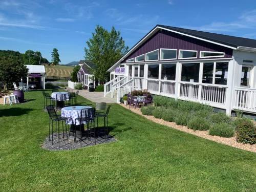 A sunny outdoor view of a restaurant with tables on a lawn, surrounded by greenery and a clear blue sky.