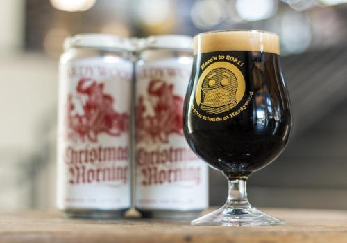 A glass of dark beer with a frothy head, next to two cans labeled "Christmas Morning."