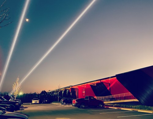 A modern building with red accents under a twilight sky, featuring beams of light and parked cars in the foreground.