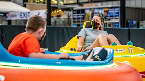 Two children wearing masks sit in colorful bumper cars, smiling and enjoying their time at an amusement venue.