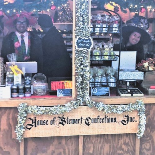 Two smiling vendors in festive hats stand behind a decorated booth labeled "House of Stewart Confections, Inc."