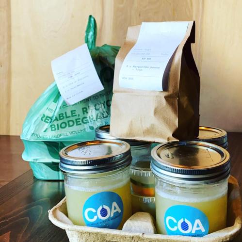 A takeout order with drinks in jars, a brown paper bag, and a green biodegradable bag on a wooden surface.