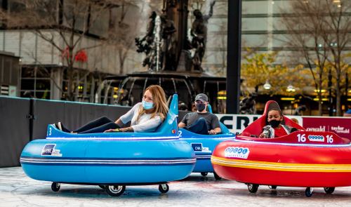 Two people in colorful bumper cars wearing masks, enjoying a fun outdoor activity in an urban setting.