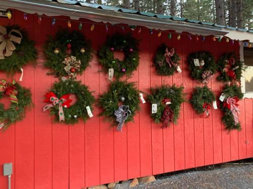 A red wall adorned with various festive wreaths, decorated with ribbons and ornaments, surrounded by holiday lights.