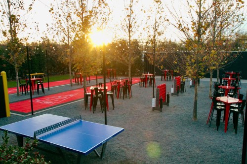 A sunlit outdoor space with tables, chairs, and a ping pong table surrounded by trees and gravel.