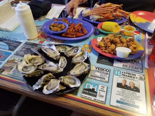 A table spread with oysters, fried shrimp, sweet potato fries, corn, and crab legs, set on a colorful menu-covered surface.