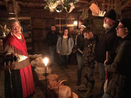 A group of people listens to a woman in historical attire, surrounded by baskets and candlelight in a rustic setting.