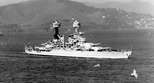 A historic battleship sails through calm waters, with mountains and a coastal town in the background. Seagulls fly nearby.