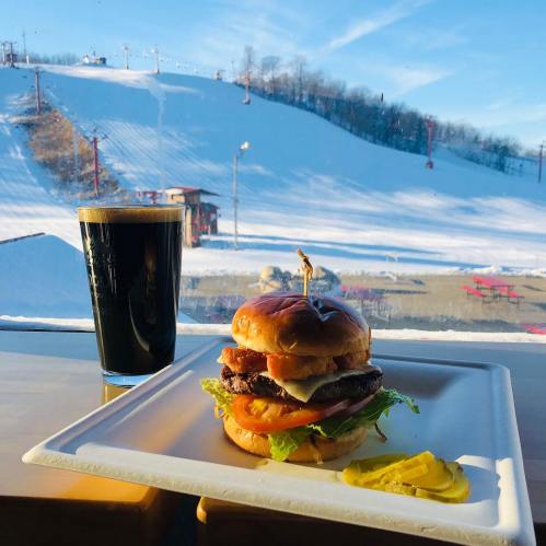 A delicious burger with lettuce, tomato, and cheese, served with a pint of dark beer, against a snowy ski slope backdrop.