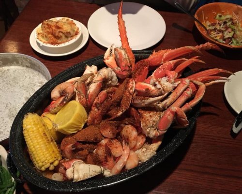 A large seafood platter with crab, shrimp, corn, and lemon, served on a dark platter with sides in the background.