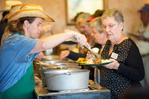 A woman in a cowboy hat serves food to a senior woman at a community meal event.