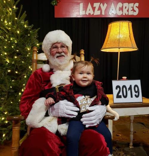 Santa Claus sits in a chair with a smiling toddler on his lap, surrounded by festive decorations and a sign reading "Lazy Acres."