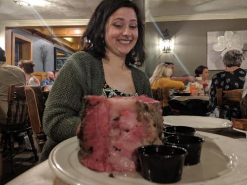A woman smiles as she sits at a table with a large cut of meat and two dipping sauces in front of her.