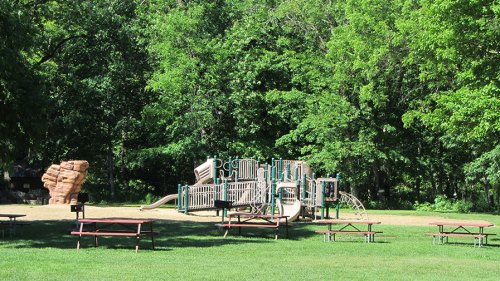 A playground with slides and climbing structures surrounded by trees, with picnic tables in the foreground.
