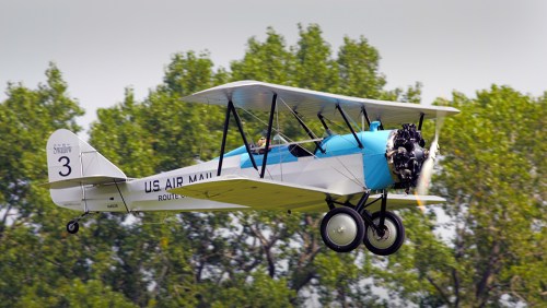 A vintage biplane with a blue and white design flying over green trees, marked "U.S. Air Mail."