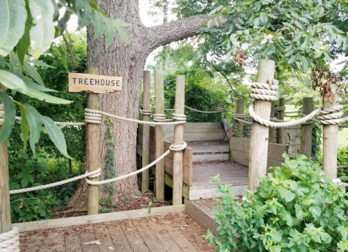 A wooden treehouse entrance with a sign, surrounded by greenery and rope railings.