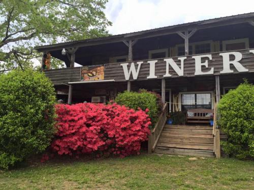 A rustic winery building with large "WINERY" sign and vibrant pink azaleas in front, surrounded by greenery.