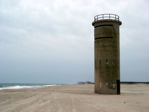A weathered concrete tower stands on a sandy beach, with a cloudy sky and ocean in the background.