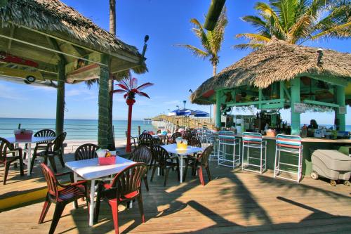 Tropical beachside bar with thatched roofs, tables, and palm trees, overlooking the ocean and a wooden pier.